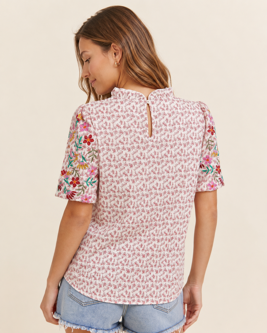 Back view of a woman wearing a pink patchwork print short-sleeve top and light wash denim shorts on a beige background