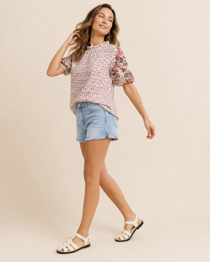 Full-body view of a woman wearing a pink patchwork print short-sleeve top, light wash denim shorts, and brown sandals on a beige background