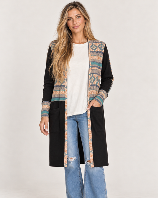 Full-body front view of a woman wearing a long black maxi cardigan with multicolor geometric patterned trim, a white top, and black wide-leg pants on a beige background.