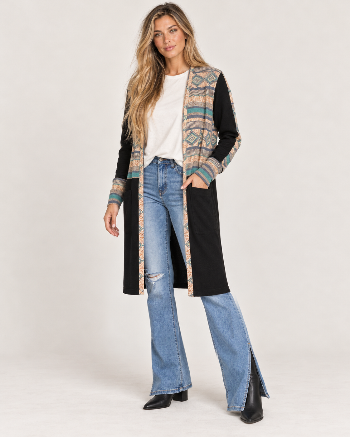 Full-body front view of a woman wearing a long black maxi cardigan with multicolor geometric patterned trim over a white top, paired with light-wash flared jeans on a beige background.
