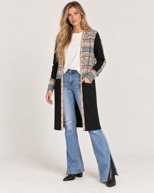 Full-body front view of a woman wearing a long black maxi cardigan with multicolor geometric patterned trim, a white top, and black wide-leg pants on a beige background.
