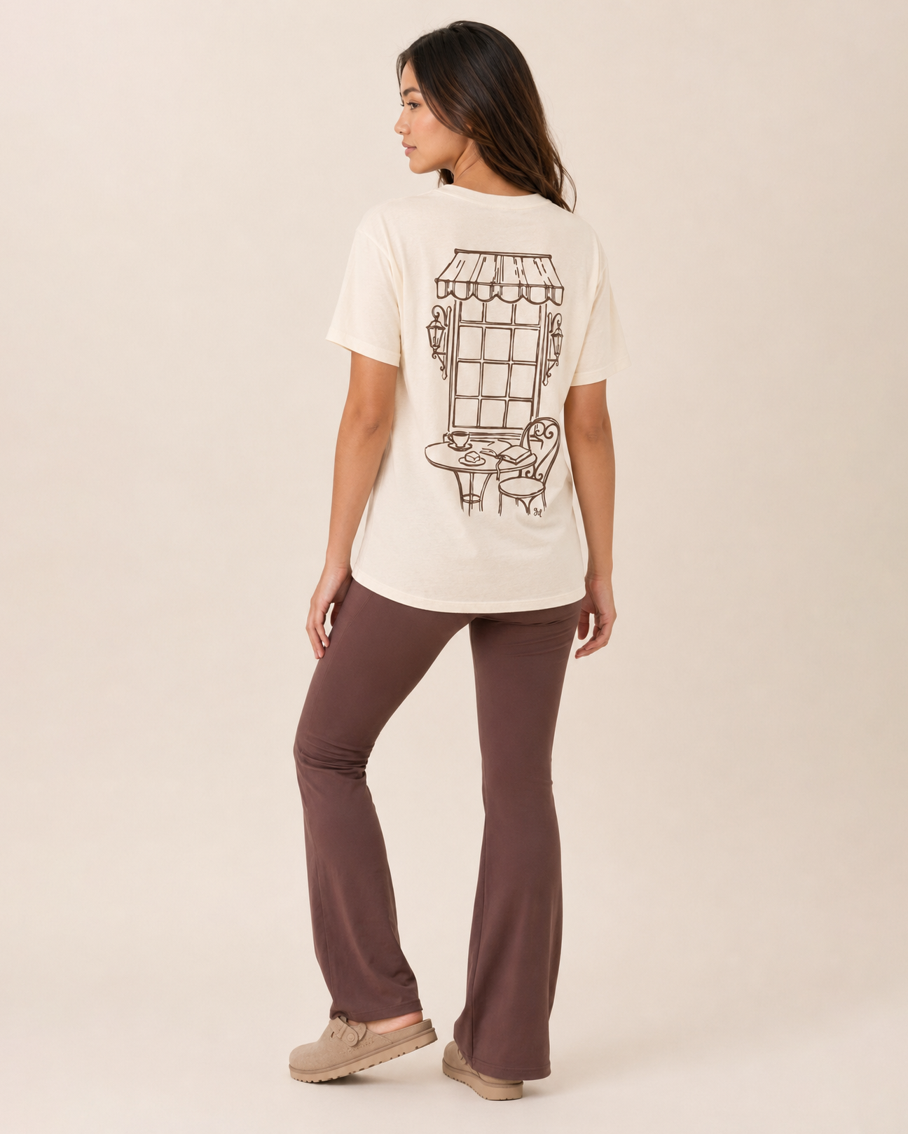 Back view of a woman wearing a cream graphic tee with a vintage-style graphic on a beige background