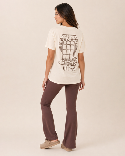 Back view of a woman wearing a cream graphic tee with a vintage-style graphic on a beige background