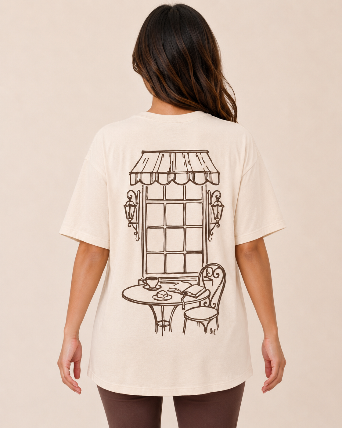 Close up back view of a woman wearing a cream graphic tee with a vintage-style graphic on a beige background