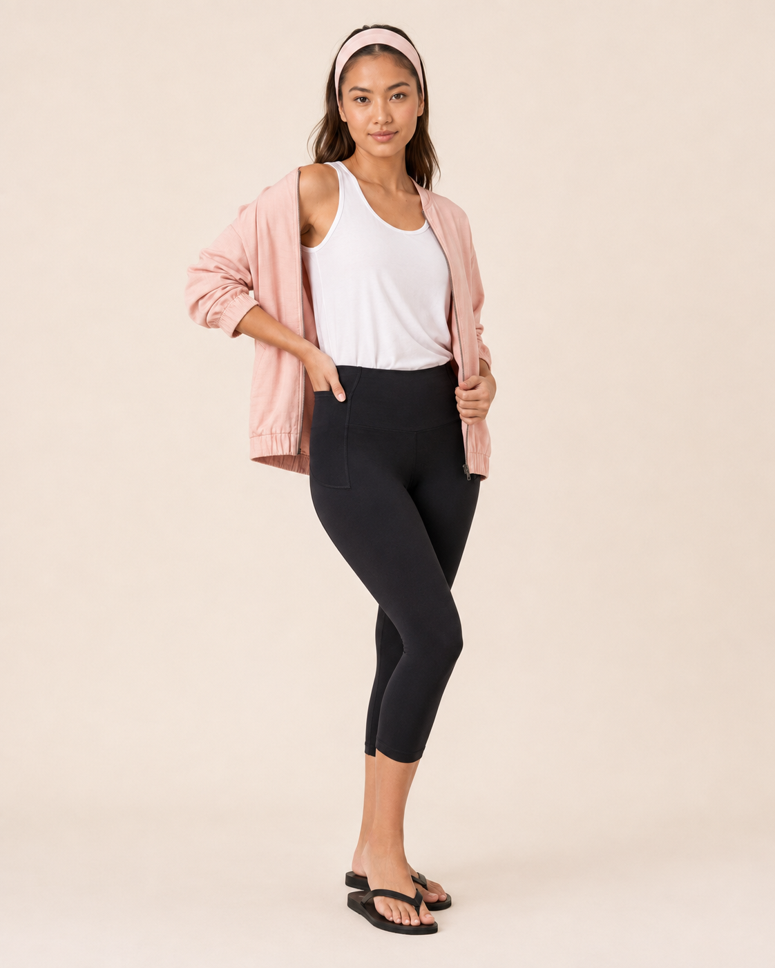 Front view of a woman wearing black capri leggings with a white top and black sandals on a beige background