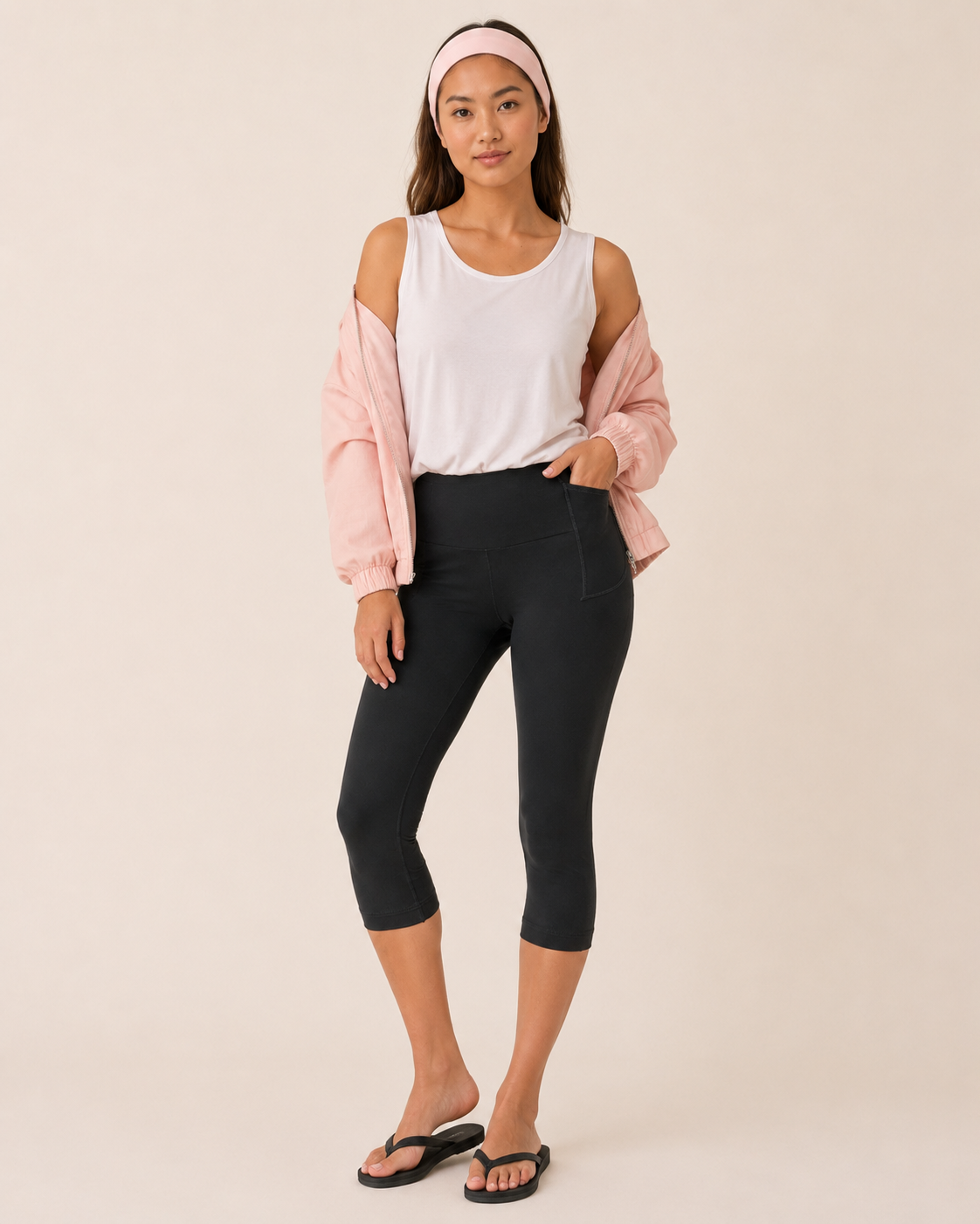 Full-body view of a woman wearing black capri leggings with a white top, pink cardigan, headband, and black sandals on a beige background