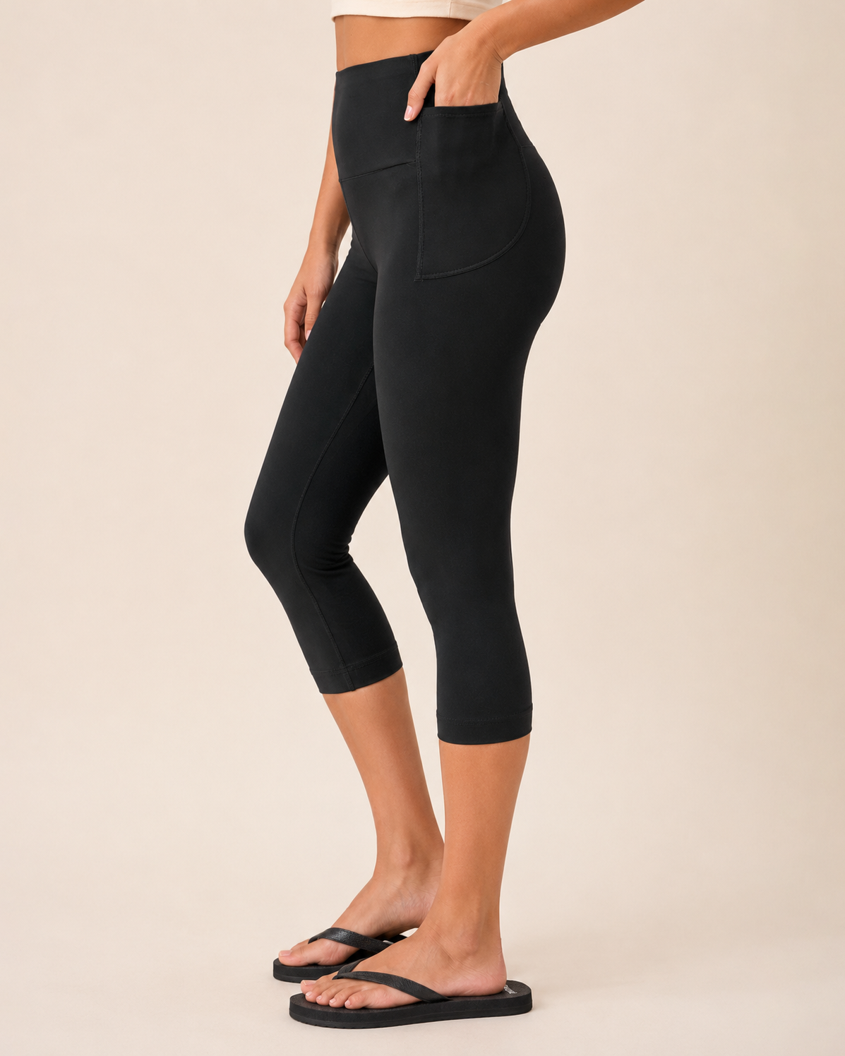 Side view of a woman wearing black capri leggings with a white top and black sandals on a beige background