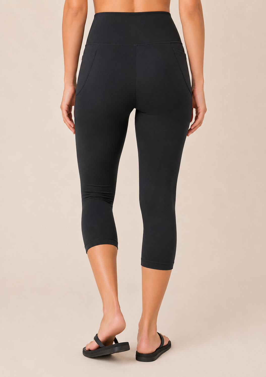 Back view of a woman wearing black capri leggings with a white top and black sandals on a beige background