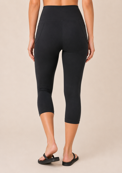 Back view of a woman wearing black capri leggings with a white top and black sandals on a beige background