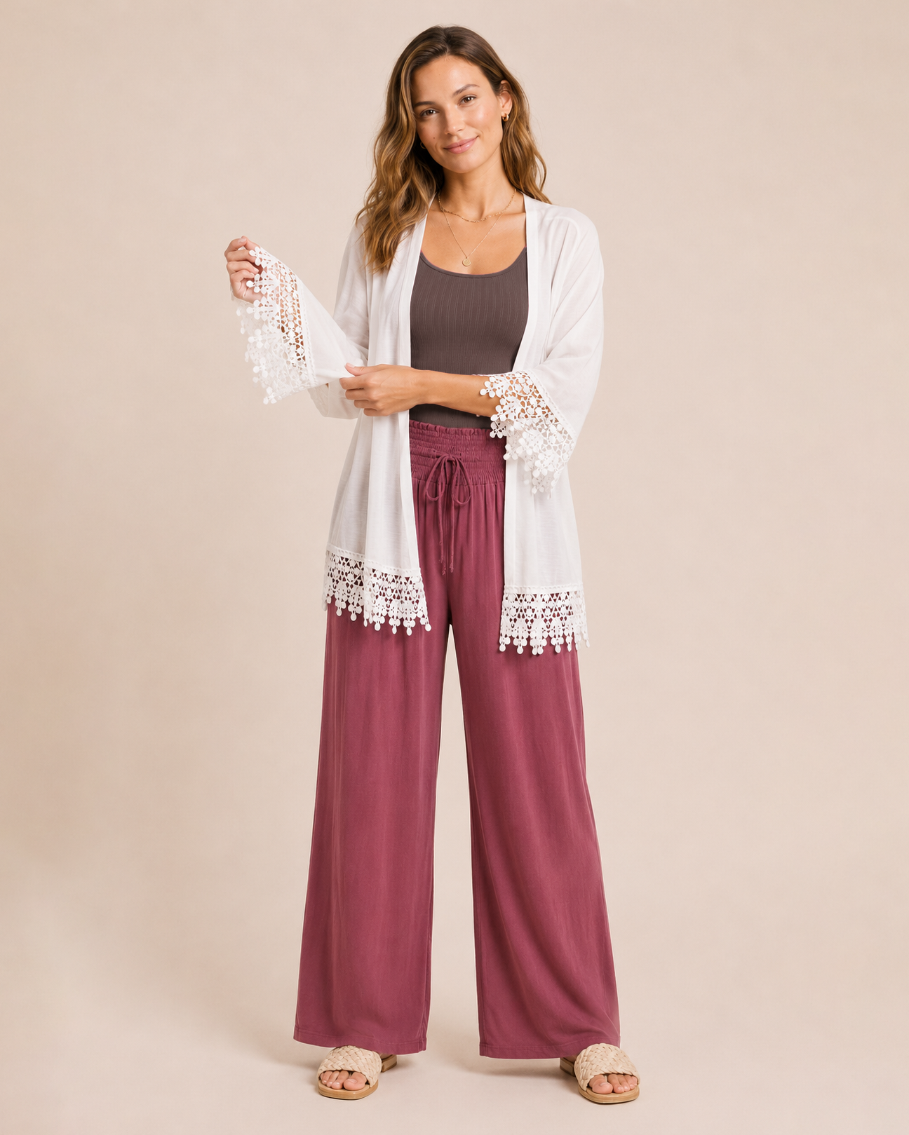 Woman wearing an ivory lace trim kimono over a brown top and burgundy wide-leg pants with tan sandals on a beige background