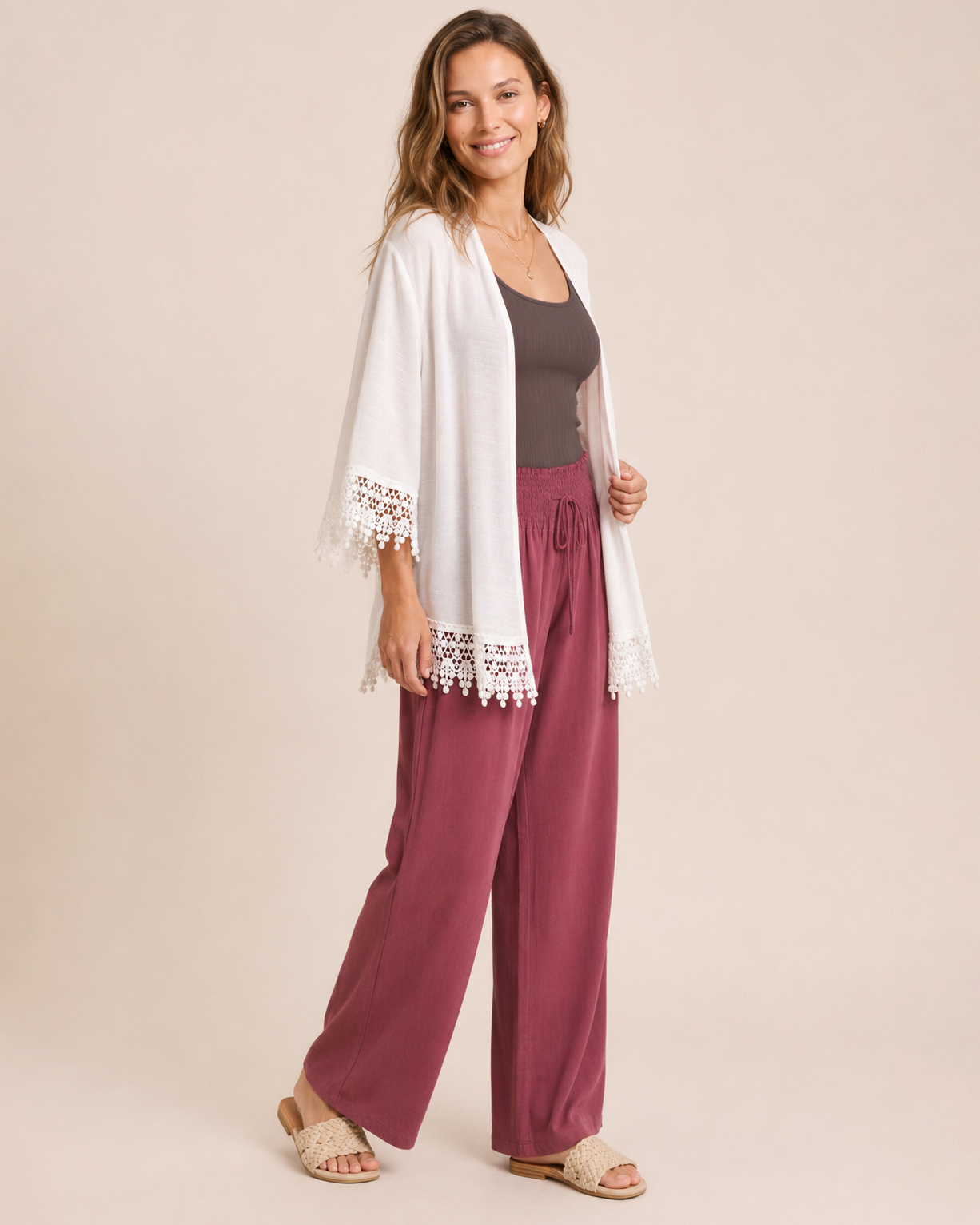 Woman wearing an ivory lace trim kimono over a brown top and burgundy wide-leg pants with tan sandals on a beige background
