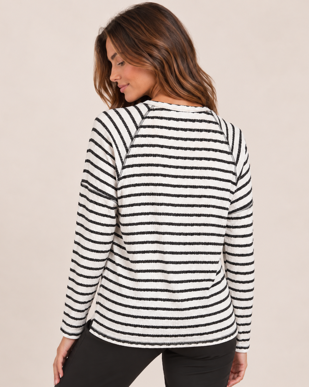 Back view of a woman wearing a black and white striped long-sleeve pullover with black pants on a beige background