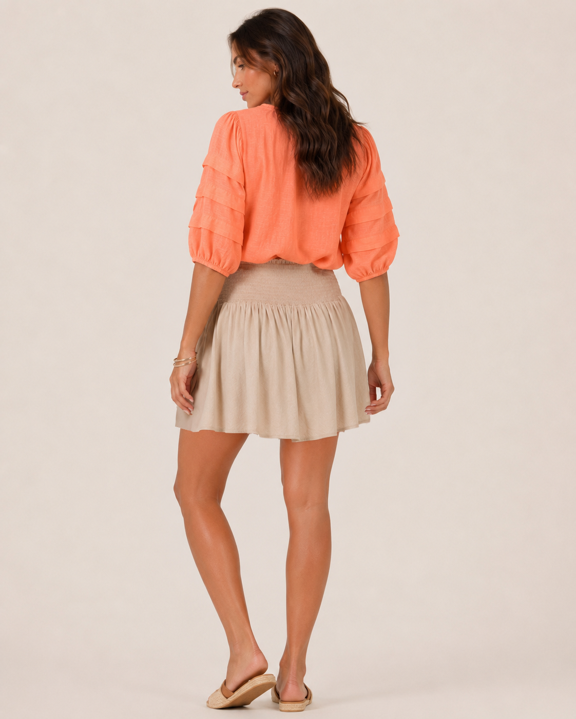 Back view of a woman wearing a sand smocked waist skort and a coral sleeveless top on a beige background