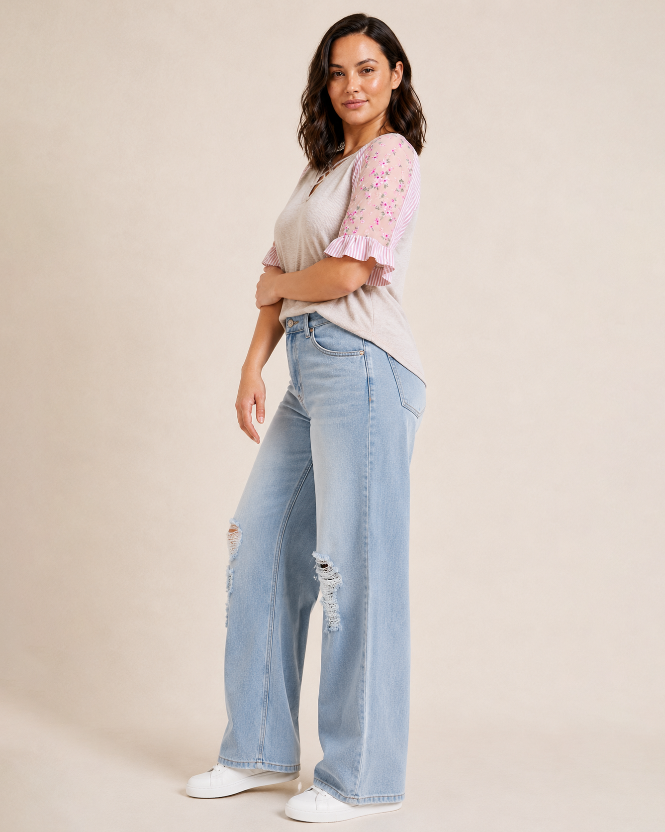 Angled full-body view of a woman wearing a blush cross-front tee with floral puff sleeves, light wash denim jeans, and beige sandals on a beige background