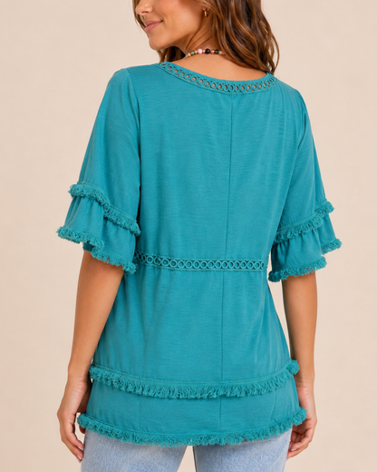 Back view of a woman wearing a teal tiered top with distressed blue jeans on a beige background