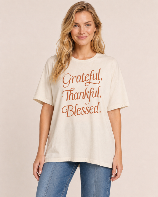 Close up view of a woman wearing a cream “Grateful, Thankful, Blessed” graphic tee with blue jeans on a beige background