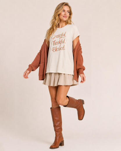 Full-body view of a woman wearing a cream “Grateful, Thankful, Blessed” graphic tee with blue jeans and tan boots on a beige background