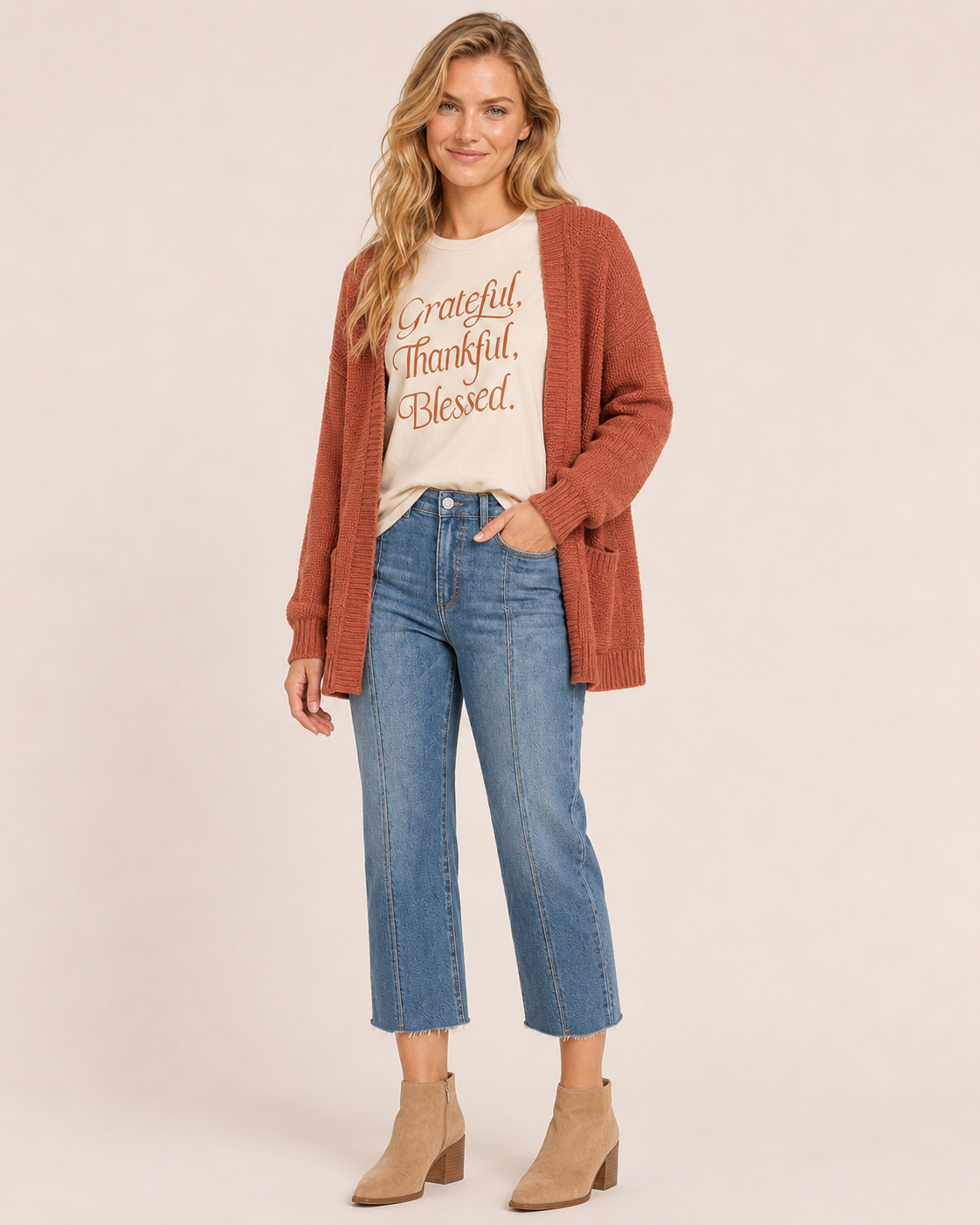 Front view of a woman wearing a cream “Grateful, Thankful, Blessed” graphic tee with blue jeans and tan boots on a beige background