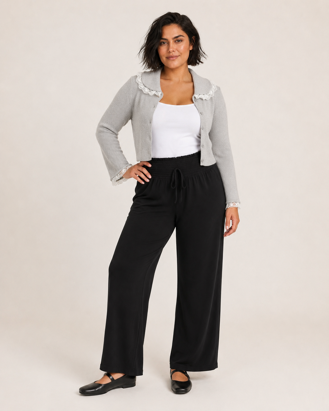 Full-body view of a woman wearing a gray lace detail flare sleeve knit cardigan with black pants and white sneakers on a beige background