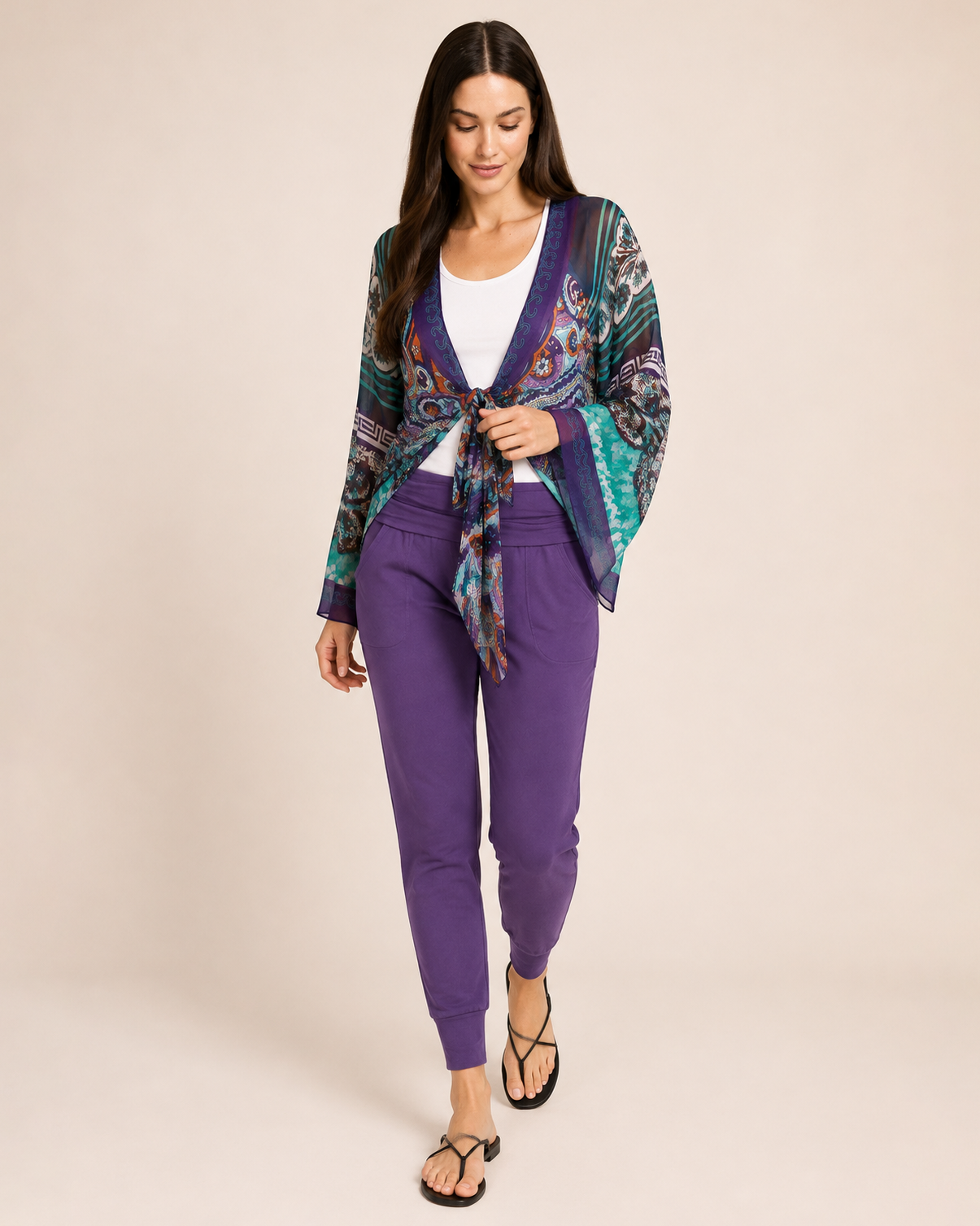Full-body view of a woman wearing eggplant foldover waist joggers with a patterned cardigan and sandals on a beige background