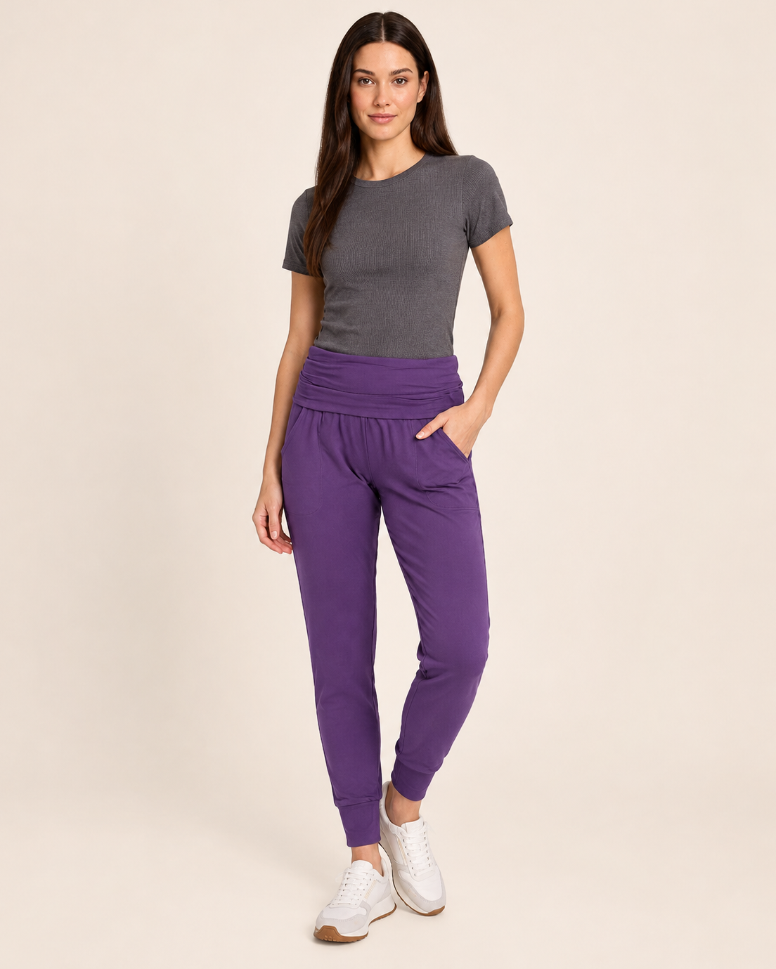 Full-body view of a woman wearing eggplant foldover waist joggers with white sneakers on a beige background