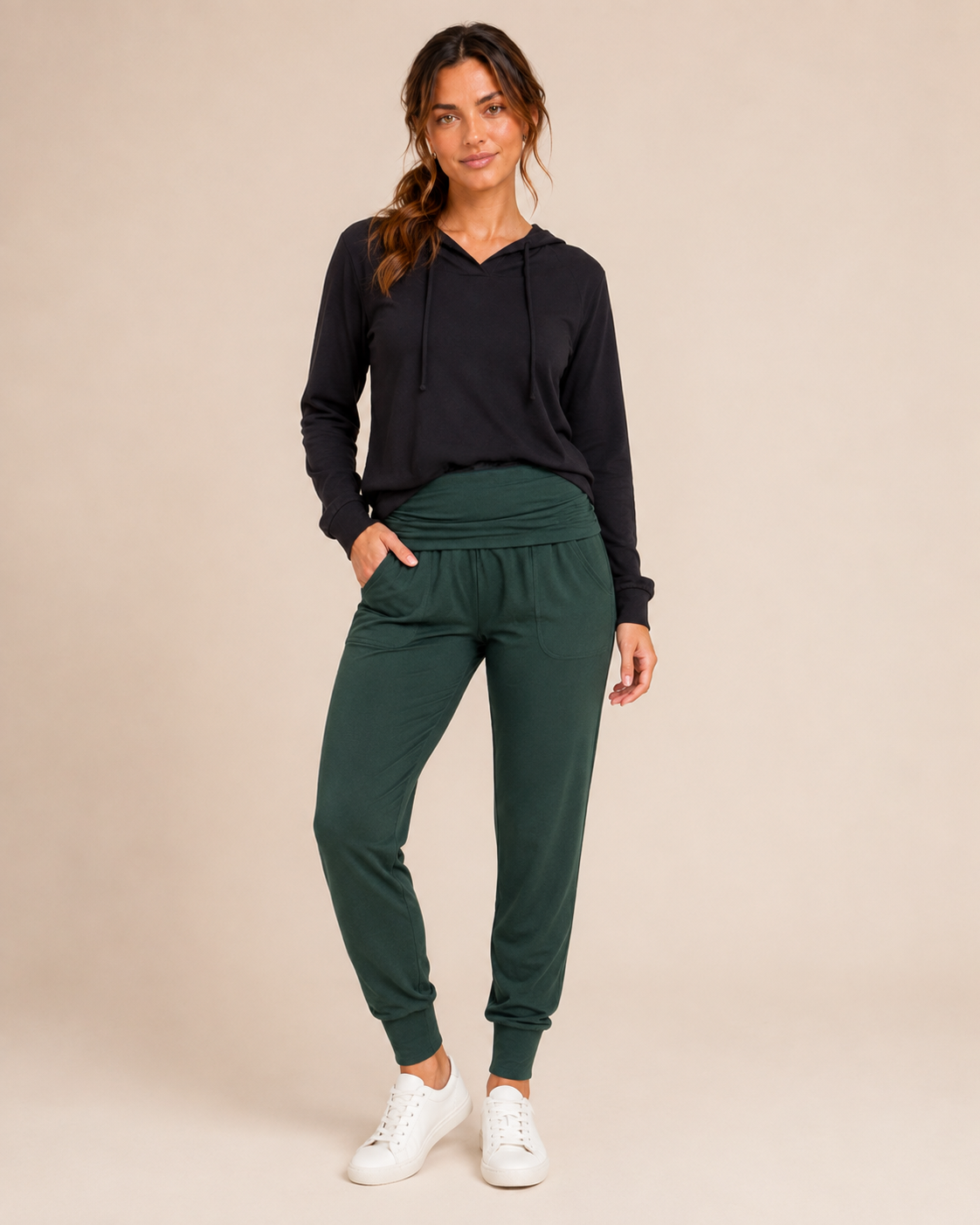 Full-body view of a woman wearing hunter green foldover waist joggers with a black top and white sneakers on a beige background