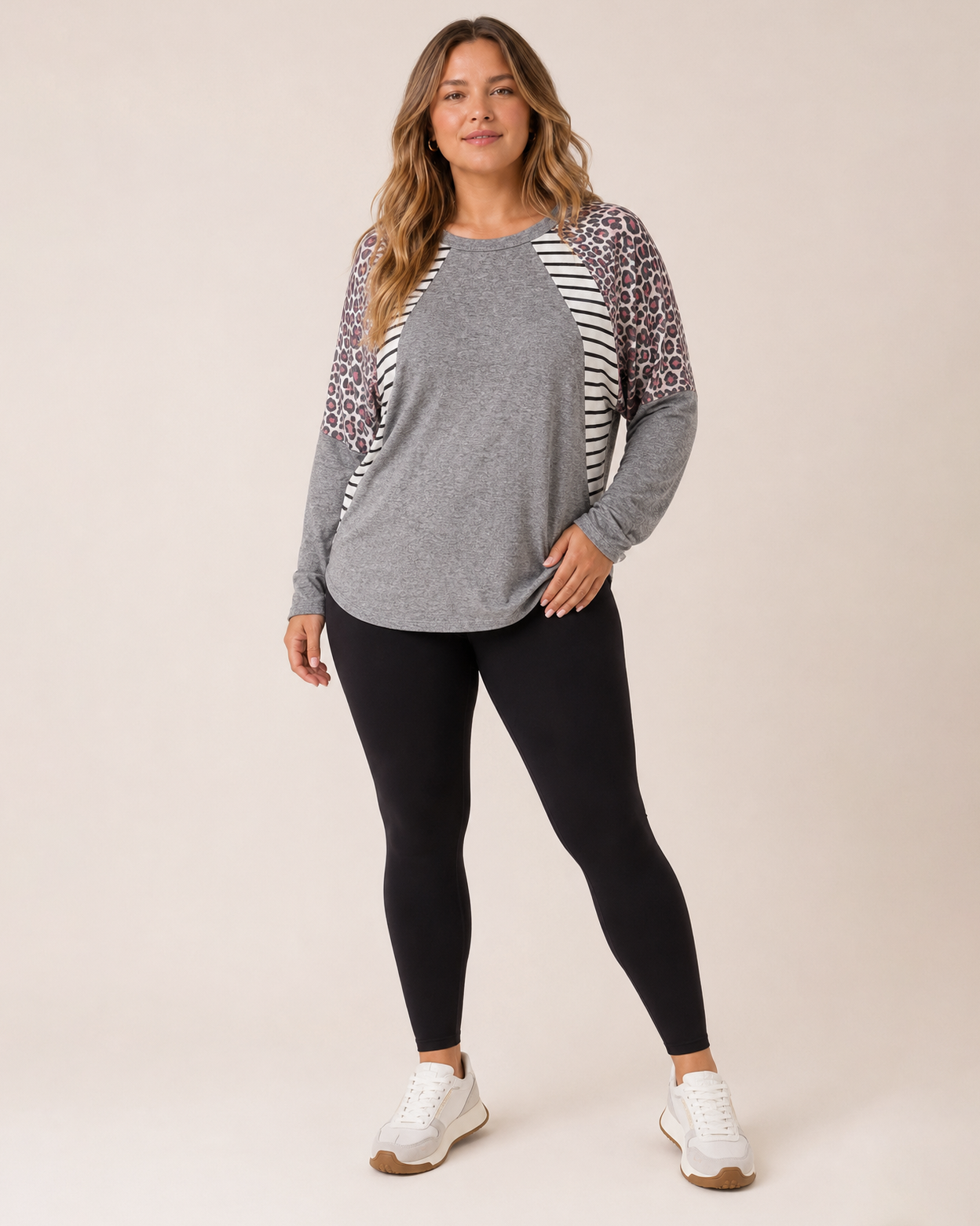 Full-body view of a woman wearing a gray oversized dolman sleeve tunic with black leggings and white sneakers on a beige background