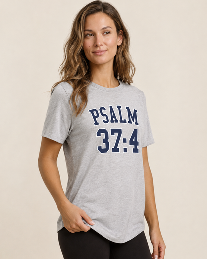 Close up view of a woman wearing a grey Psalm 37:4 graphic tee with a blue text design on a beige background