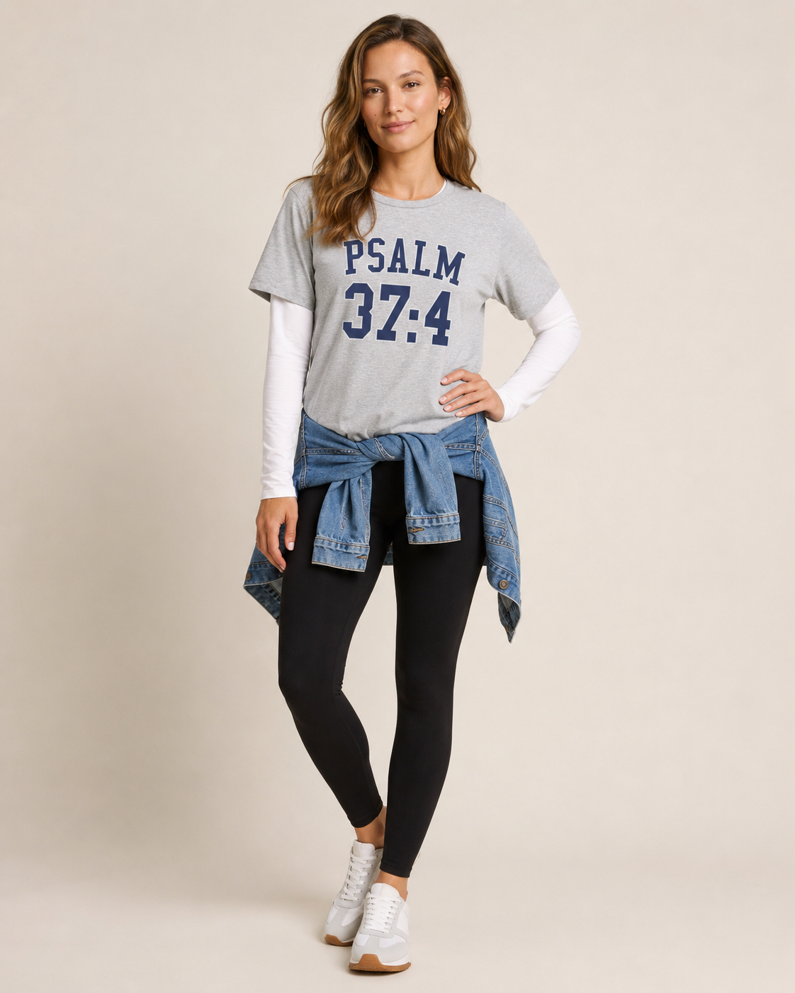 Full-body view of a woman wearing a grey Psalm 37:4 graphic tee with black pants and white sneakers on a beige background
