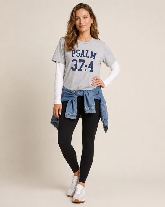 Close up view of a woman wearing a grey Psalm 37:4 graphic tee with a blue text design on a beige background