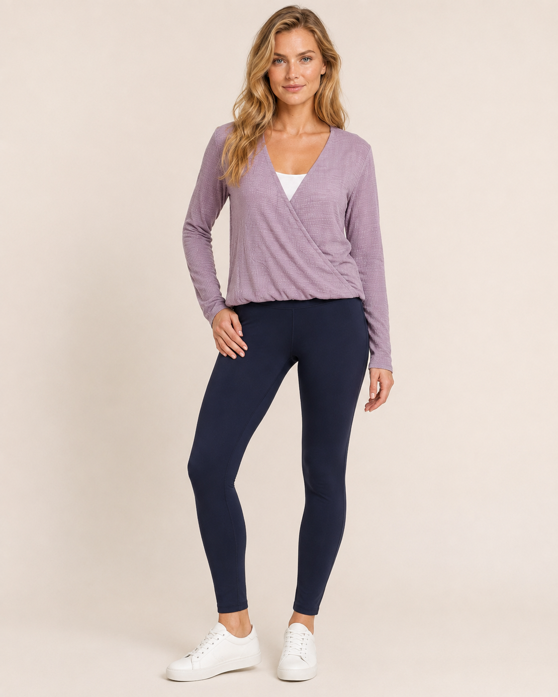 Full-body view of a woman wearing a lilac crossover blouse with black pants and white sneakers on a beige background