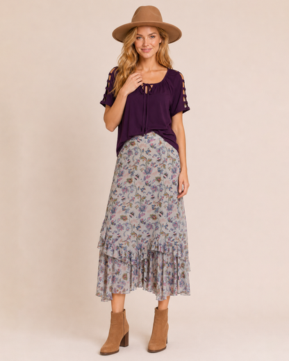 Full-body view of a woman wearing a purple ruffle-sleeve top with a floral midi skirt, brown ankle boots, and a brown felt hat on a beige background