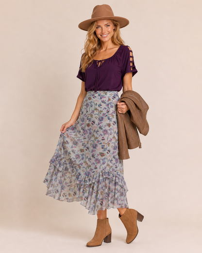 Side view of a woman wearing a purple ruffle-sleeve top with a floral midi skirt, brown ankle boots, and a brown felt hat on a beige background