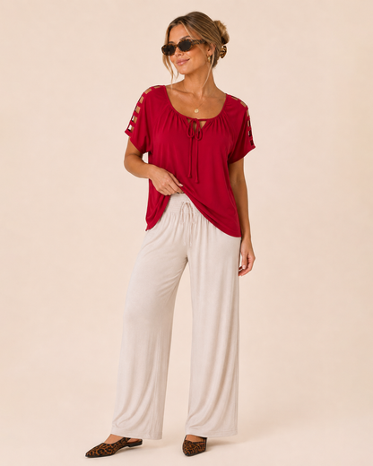 Side view of a woman wearing a red short-sleeve top and black glasses with white wide-leg pants and leopard-print flats on a beige background