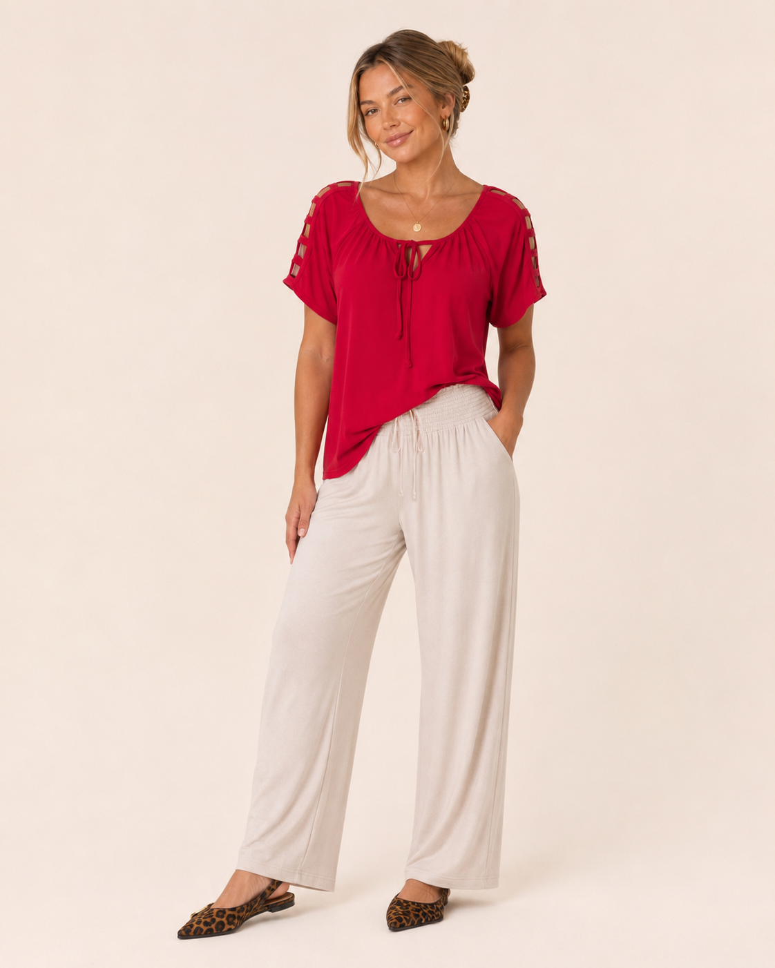 Full-body view of a woman wearing a red short-sleeve top with white wide-leg pants and leopard-print flats on a beige background