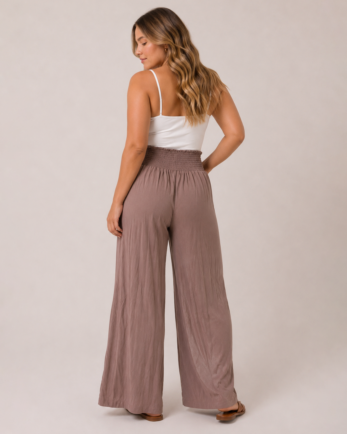 Back view of a woman wearing mocha smocked waist wide-leg pants and a white tank top on a beige background
