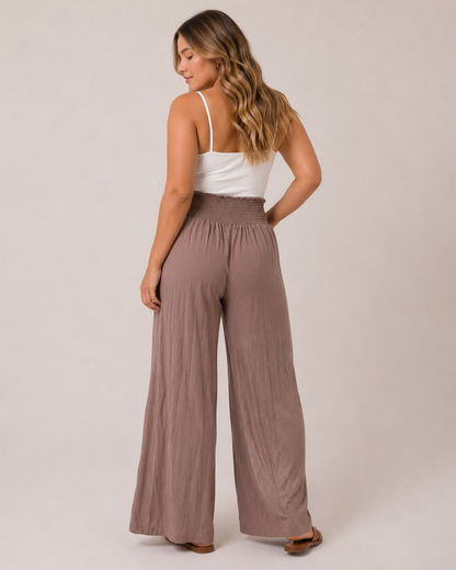 Back view of a woman wearing mocha smocked waist wide-leg pants and a white tank top on a beige background
