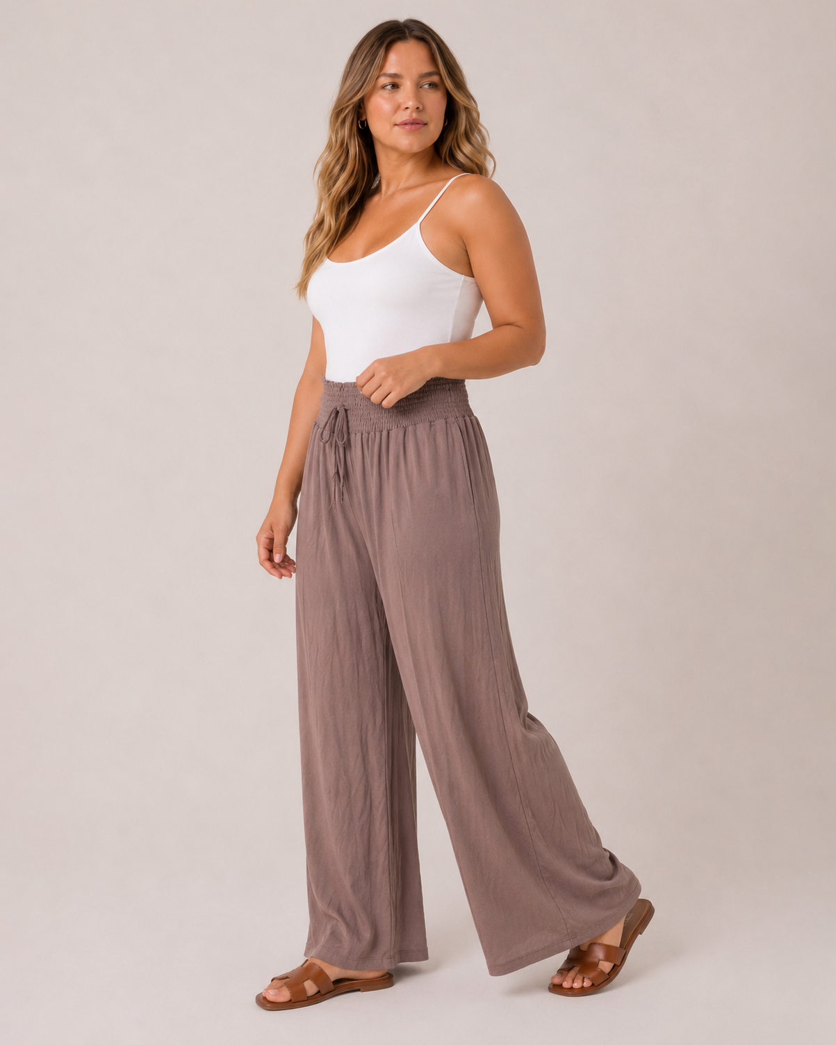 Front view of a woman wearing mocha smocked waist wide-leg pants and a white tank top on a beige background