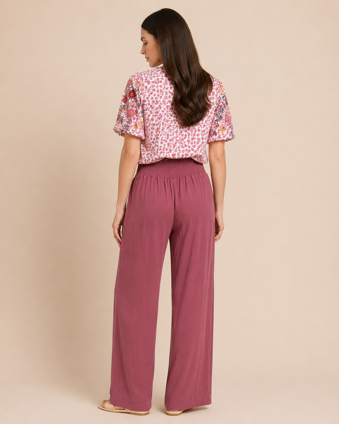 Back view of a woman wearing rosewood smocked waist wide-leg pants and a pink floral blouse on a beige background