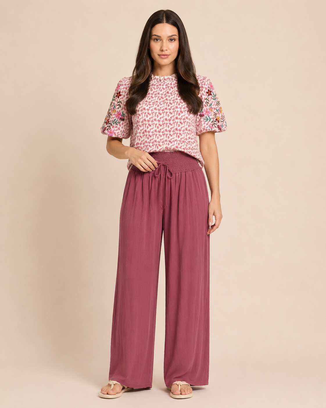 Front view of a woman wearing rosewood smocked waist wide-leg pants and a pink floral blouse on a beige background