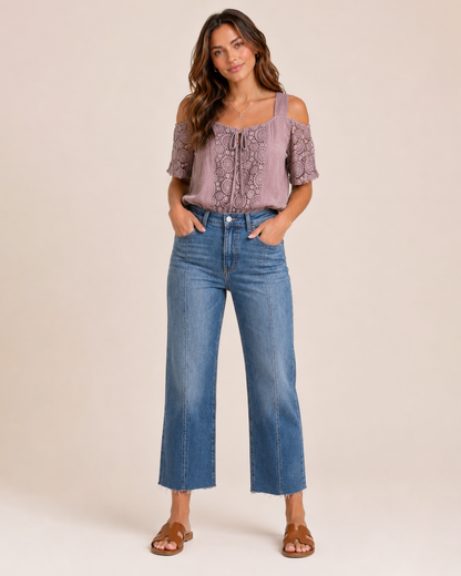 Full-body view of a woman wearing a mauve cold shoulder lace blouse, cropped blue jeans, and brown sandals on a beige background