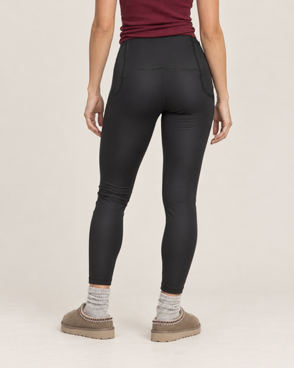 Cropped back view of a woman wearing black high-waisted leggings, showing the waistband, fitted seat, and ankle-length legs on a beige background.