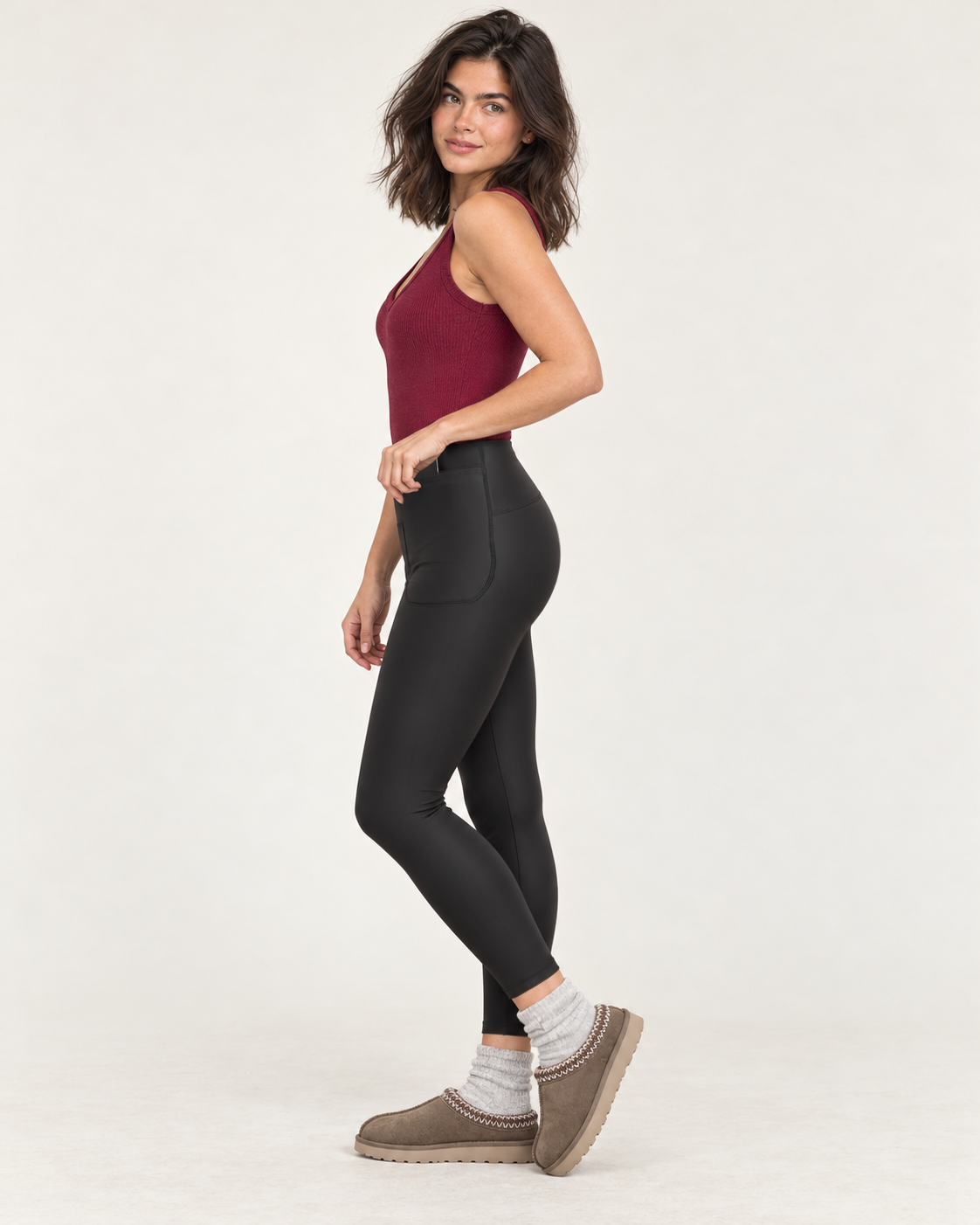 Full-body side view of a woman wearing black high-waisted leggings with a burgundy tank top and taupe sneakers on a beige background.