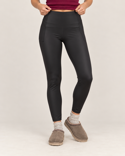 Full-body front view of a woman wearing black high-waisted sculpting leggings with a burgundy tank top and taupe sneakers on a beige background.