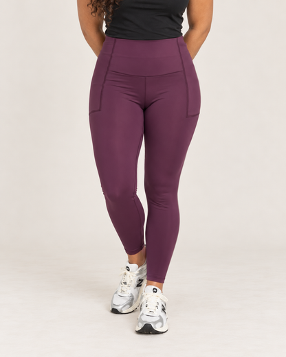 Cropped front view of a woman wearing high-waisted plum leggings with a black sleeveless top, highlighting the wide waistband and smooth, fitted legs against a beige background.