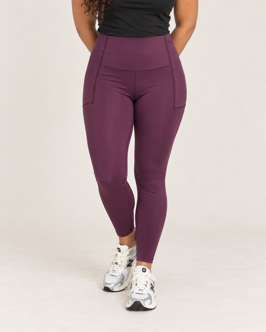 Front view of a woman wearing high-waisted plum leggings with a black sleeveless top and black open-front cardigan against a beige background.