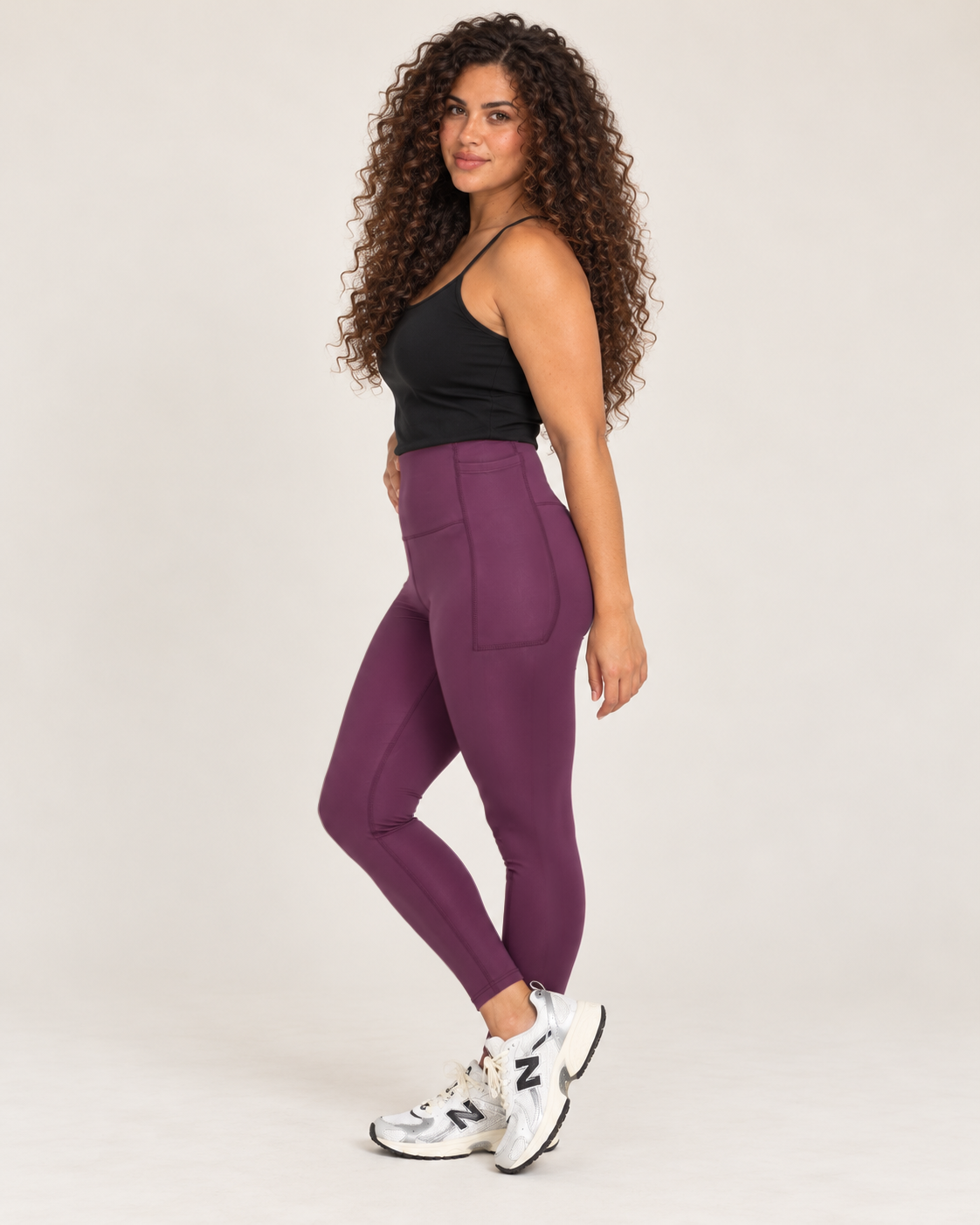Side view of a woman wearing high-waisted plum leggings with a black sleeveless top, showcasing the fitted silhouette and full-length design against a beige background.