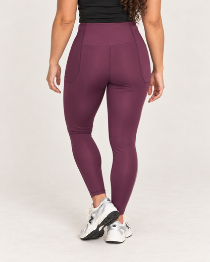Back view of a woman wearing high-waisted plum leggings with a fitted waistband and full-length legs, highlighting the smooth back and contoured fit against a beige background.