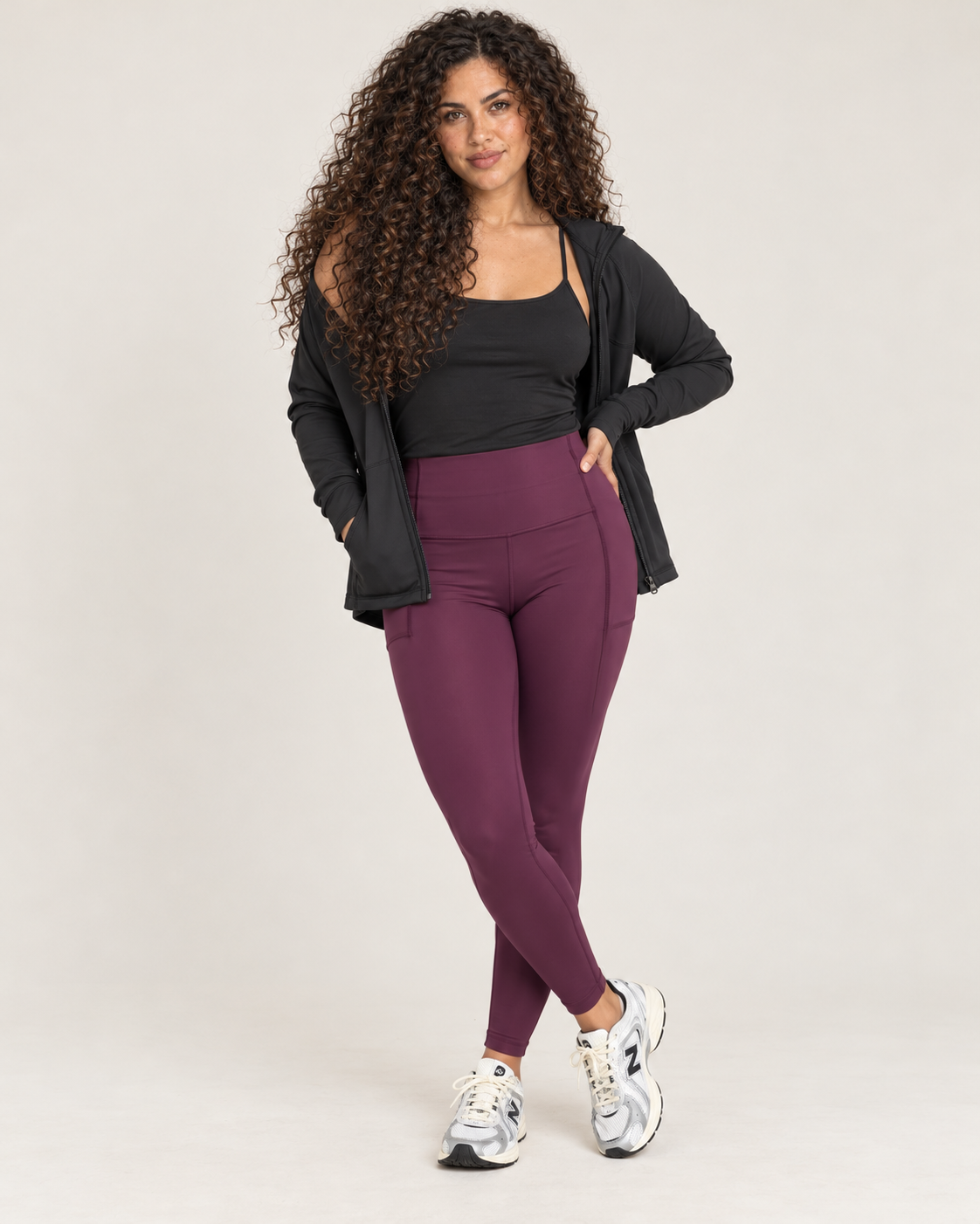 Front view of a woman wearing high-waisted plum leggings with a black sleeveless top and black open-front cardigan against a beige background.