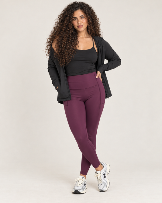 Front view of a woman wearing high-waisted plum leggings with a black sleeveless top and black open-front cardigan against a beige background.